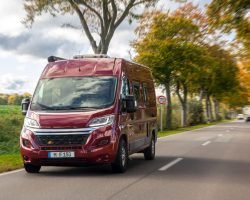 Red camper van driving on a countryside road surrounded by trees and fields in autumn. Symbol of freedom, travel, and modern lifestyle adventure.